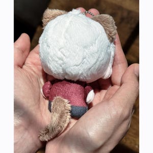 May include: A plush keychain in the shape of a small animal, featuring a white head, brown ears, and a fluffy tail. The animal wears a burgundy outfit with gray pants. A silver chain is attached to the top of the plush toy.