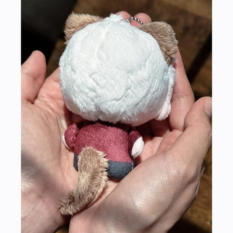 May include: A plush keychain in the shape of a small animal, featuring a white head, brown ears, and a fluffy tail. The animal wears a burgundy outfit with gray pants. A silver chain is attached to the top of the plush toy.