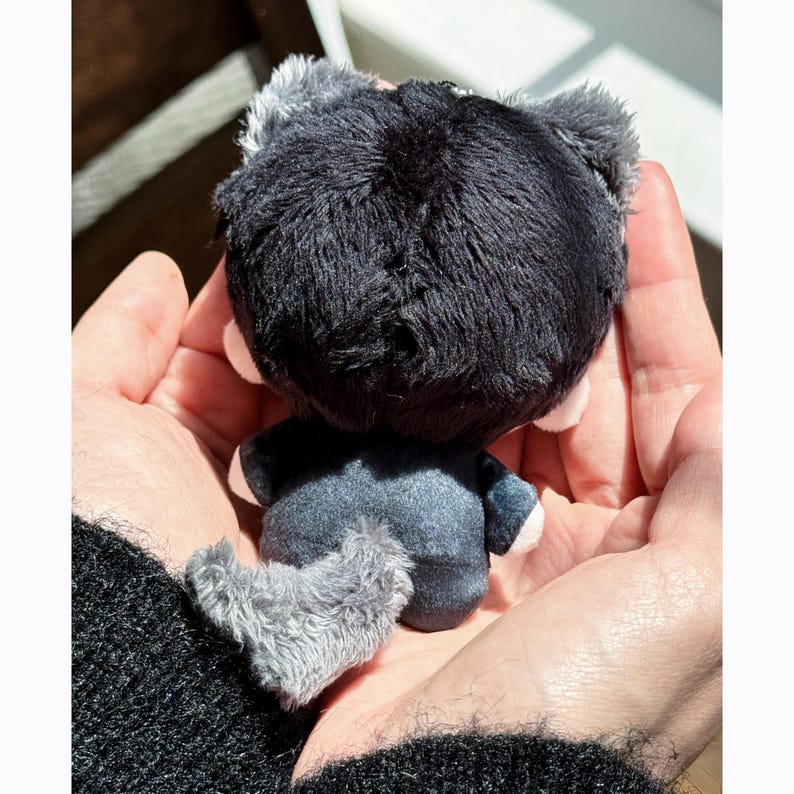 May include: A small, plush toy in the shape of a wolf, held in open hands. The wolf has a dark gray body, a fluffy black head, and a gray tail. The toy's ears are also gray. The wolf is wearing a dark gray outfit.