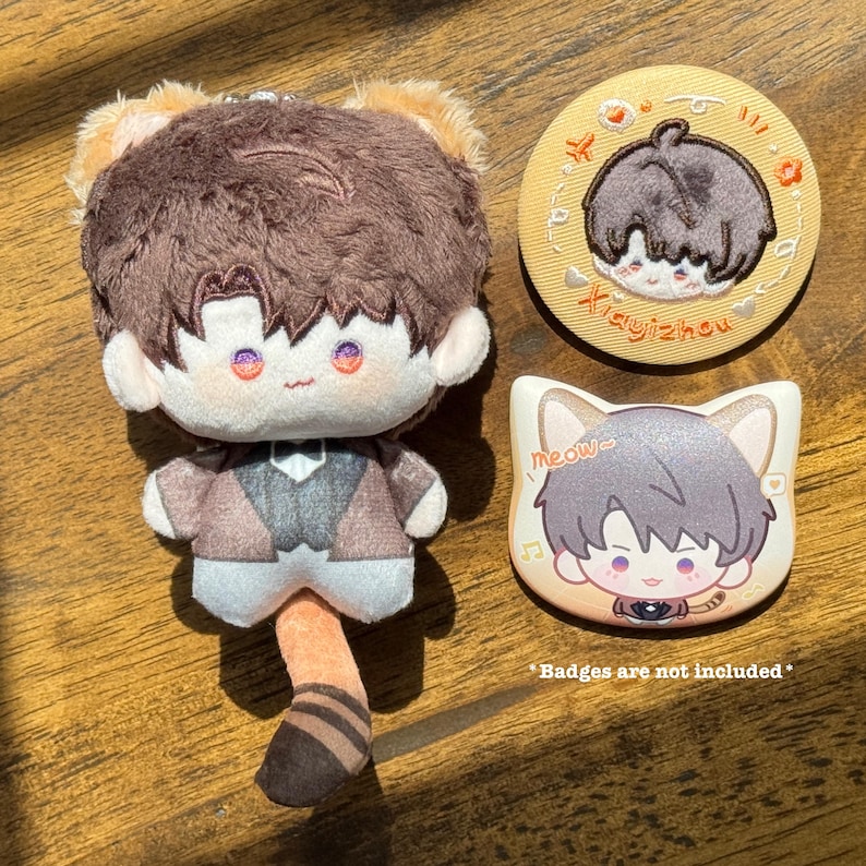 May include: A plush doll with brown hair and cat ears, wearing a brown jacket and vest. Two decorative badges are also present, one round with text and the other cat-shaped with the word "meow".