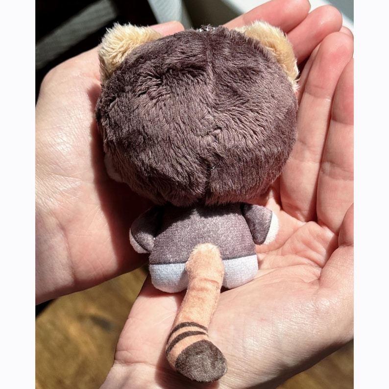 May include: A plush toy in the shape of a brown bear, viewed from the back. The bear has a round head with tan ears, a gray body, and a long, striped tail. The toy is held in open hands.