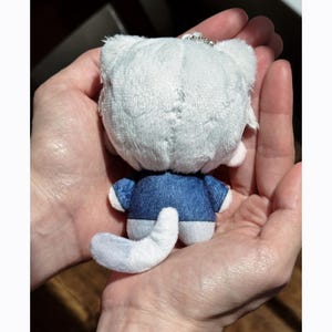 May include: A small, plush cat keychain in shades of white and blue. The cat has a white head and tail, with a blue jacket and white accents. The keychain is held in open hands.