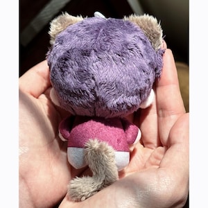 May include: A plush toy cat with a purple body, pink shirt, and a gray tail. The cat has gray ears and white accents. The toy is held in open hands, showcasing its soft texture and small size. The cat is a cute and cuddly toy.