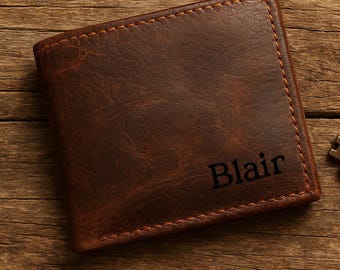 May include: A brown leather wallet with the name "Blair" embossed on the front, a vintage-style brass key, and a matching leather keychain. The items are arranged on a wooden surface.