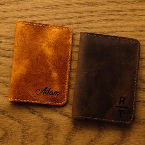 May include: Two leather wallets, one tan and one brown, rest on a wooden surface. The tan wallet is embossed with the name "Adam". The brown wallet features the initials "R" and "T". Both wallets have stitched edges.