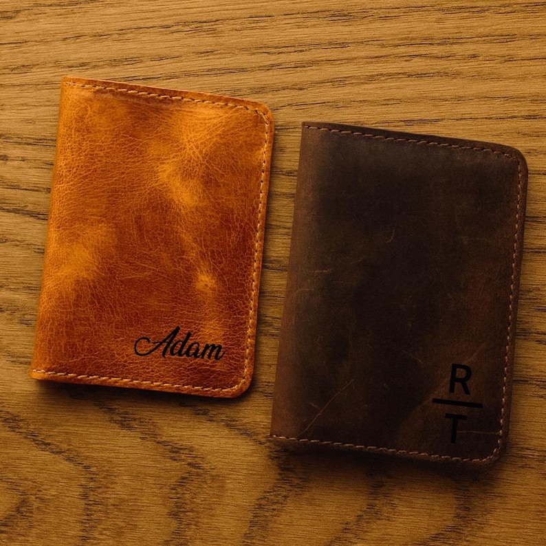 May include: Two leather wallets, one tan and one brown, rest on a wooden surface. The tan wallet is embossed with the name "Adam". The brown wallet features the initials "R" and "T". Both wallets have stitched edges.