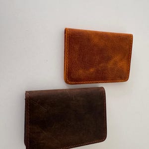 May include: Two leather cardholders in shades of brown. The top cardholder is a warm, reddish-brown, while the bottom one is a darker, chocolate brown. Both have a rectangular shape with rounded corners and visible stitching.