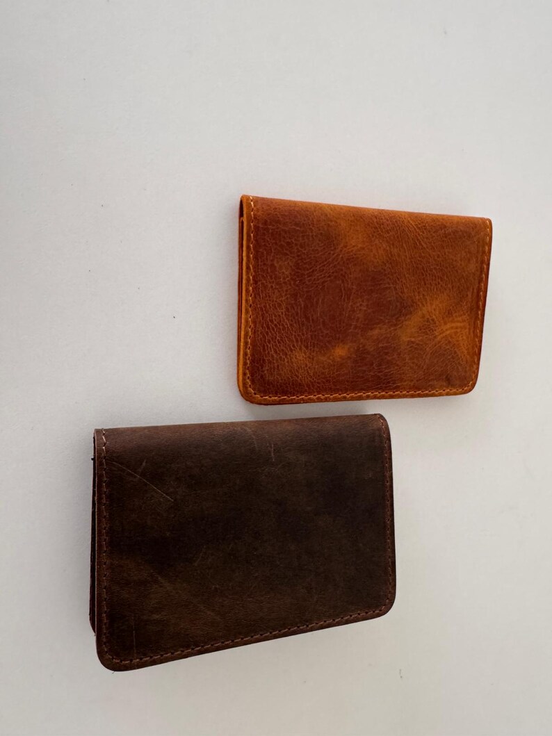 May include: Two leather cardholders in shades of brown. The top cardholder is a warm, reddish-brown, while the bottom one is a darker, chocolate brown. Both have a rectangular shape with rounded corners and visible stitching.
