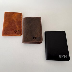 May include: Three leather wallets in shades of brown and black. The wallets are rectangular and feature personalized initials: "GP", "Blair", and "SFH". The wallets are likely designed for carrying cards and cash.
