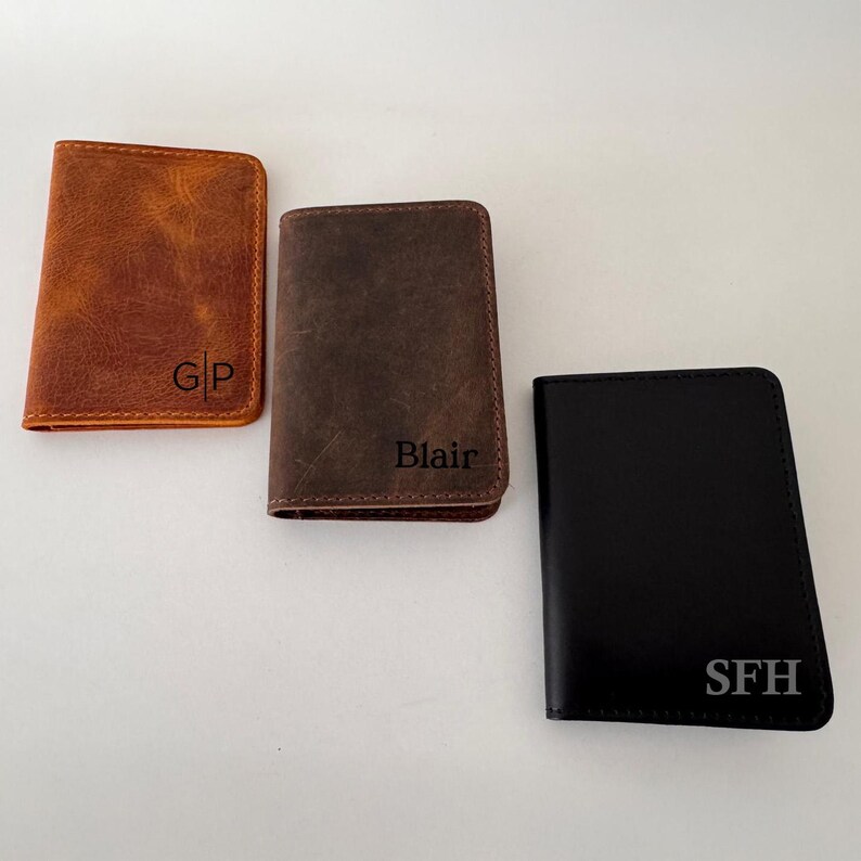 May include: Three leather wallets in shades of brown and black. The wallets are rectangular and feature personalized initials: "GP", "Blair", and "SFH". The wallets are likely designed for carrying cards and cash.
