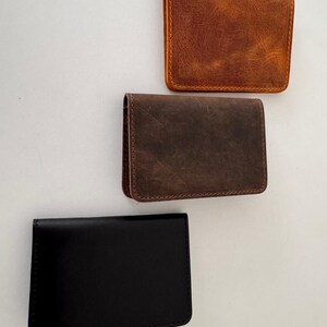 May include: Three leather card holders in different colors. The top one is a distressed brown, the middle one is a darker brown, and the bottom one is black. All have a rectangular shape with rounded corners.