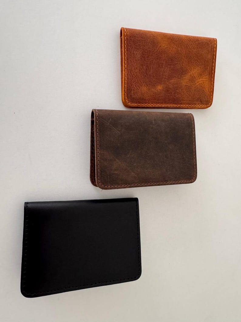 May include: Three leather card holders in different colors. The top one is a distressed brown, the middle one is a darker brown, and the bottom one is black. All have a rectangular shape with rounded corners.
