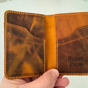 May include: Open leather wallet with two card slots and the words "Super Papa" embossed on the interior. The wallet is brown with a distressed finish and yellow stitching.