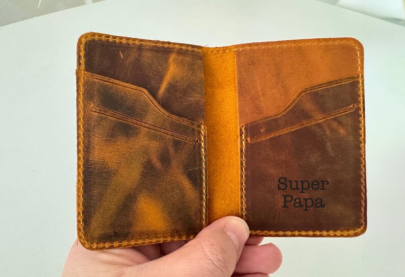 May include: Open leather wallet with two card slots and the words "Super Papa" embossed on the interior. The wallet is brown with a distressed finish and yellow stitching.