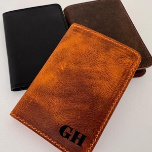 May include: Three leather wallets in black, brown, and tan. The tan wallet has the initials "GH" in black. The wallets are rectangular and appear to be made of genuine leather. The wallets are likely for carrying cards and cash.