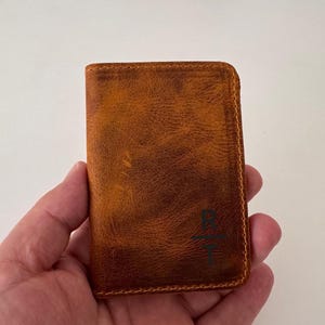 May include: A brown leather wallet with a textured surface. The wallet has a rectangular shape and is held in a hand. The wallet has a stitched border and a dark green graphic on the front.
