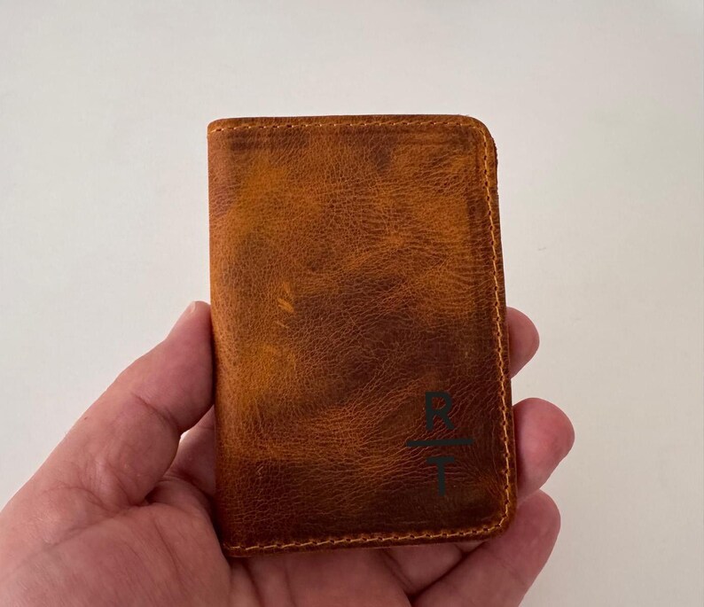 May include: A brown leather wallet with a textured surface. The wallet has a rectangular shape and is held in a hand. The wallet has a stitched border and a dark green graphic on the front.