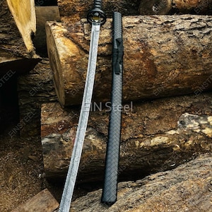 May include: A katana sword with a patterned Damascus steel blade, a black wrapped handle, and a circular guard. Beside the blade lies a matching black scabbard featuring a textured diamond pattern and a black cord wrap detail near the top.