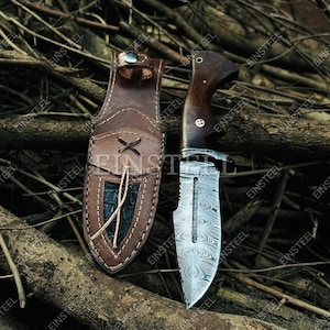 Pu&ograve; includere: Un coltello a lama fissa con lama in acciaio damasco a motivi e manico in legno scuro, dotato di una guardia in metallo e un perno decorativo. Accanto si trova un fodero in pelle marrone con bordo cucito, dettaglio a lacci e inserto scuro.