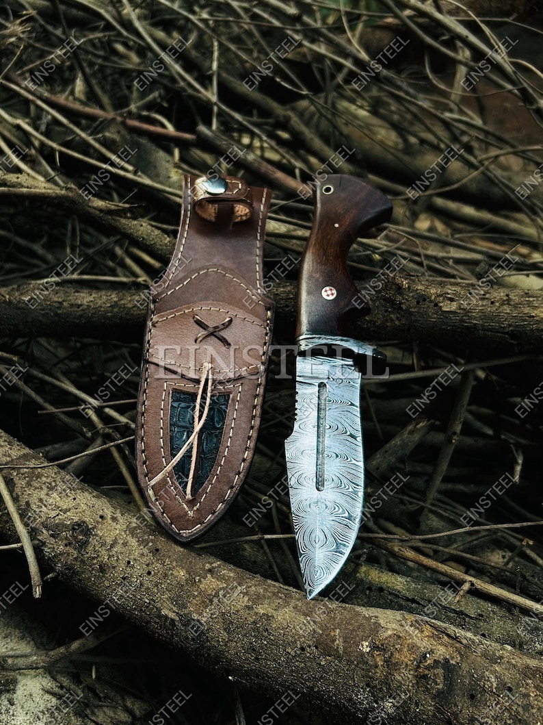 Pu&ograve; includere: Un coltello a lama fissa con lama in acciaio damasco a motivi e manico in legno scuro, dotato di una guardia in metallo e un perno decorativo. Accanto si trova un fodero in pelle marrone con bordo cucito, dettaglio a lacci e inserto scuro.