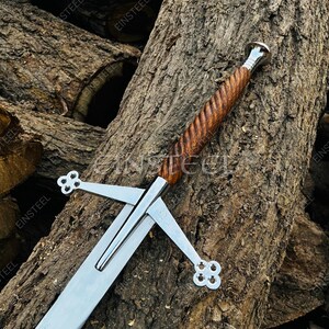 May include: A Scottish claymore sword with a polished steel blade and a wooden handle. The handle is intricately carved with a spiral design. The crossguard features a quatrefoil design. The sword is displayed against a backdrop of rough-hewn logs.