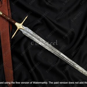 May include: A long, ornate sword with a patterned blade and a gold-colored crossguard and handle. The handle is dark brown, and the sword rests on a black, textured surface. A brown leather sheath is visible.