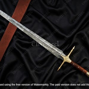 May include: A long, ornate sword with a patterned blade, gold crossguard, and a dark wooden handle. The sword rests on a black fabric with a brown leather strap. The blade has a length of approximately 30 inches.