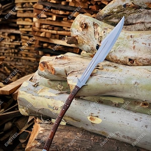 May include: A spear with a long, polished silver blade and a dark brown wooden handle. The handle features intricate carved designs. The spear is resting on a pile of logs, with a background of more logs.
