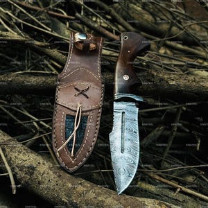 May include: A handcrafted hunting knife with a patterned steel blade and a dark wooden handle. The knife is displayed with a brown leather sheath, featuring decorative stitching and a textured inlay. The background is a natural setting with branches.