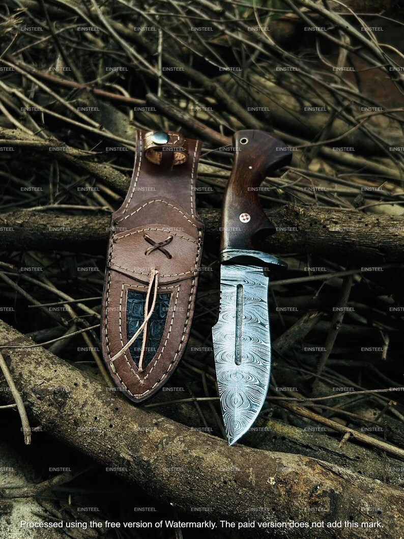 May include: A handcrafted hunting knife with a patterned steel blade and a dark wooden handle. The knife is displayed with a brown leather sheath, featuring decorative stitching and a textured inlay. The background is a natural setting with branches.