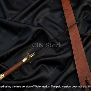 May include: A sword with a dark blade, wooden handle, and gold-colored accents. The sword rests on a black fabric surface, accompanied by a brown leather sheath. The text "EIN Steel" is visible.