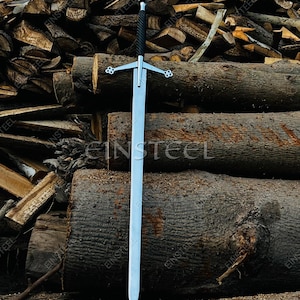 May include: A long, silver sword with a black handle and cross-shaped hilt. The sword is displayed vertically against a backdrop of stacked logs. The blade is long and straight, with a pointed tip. The hilt features decorative elements.