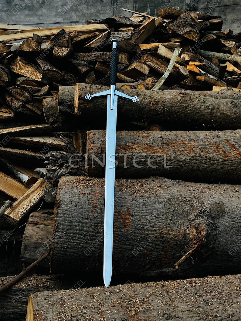 May include: A long, silver sword with a black handle and cross-shaped hilt. The sword is displayed vertically against a backdrop of stacked logs. The blade is long and straight, with a pointed tip. The hilt features decorative elements.