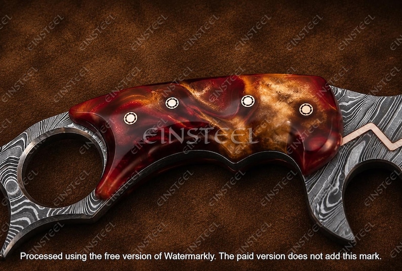 Pu&ograve; includere: Un manico di coltello con un'impugnatura in resina lucida dai motivi rossi e ambrati, fissata da tre rivetti metallici circolari. La lama presenta un distinto motivo in acciaio Damasco grigio e nero con un dettaglio in rame intarsiato.