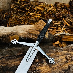May include: A medieval-style sword with a polished steel blade and crossguard. The handle is wrapped in black material, and the pommel is silver. The sword rests on a weathered wooden surface, with a background of stacked firewood.