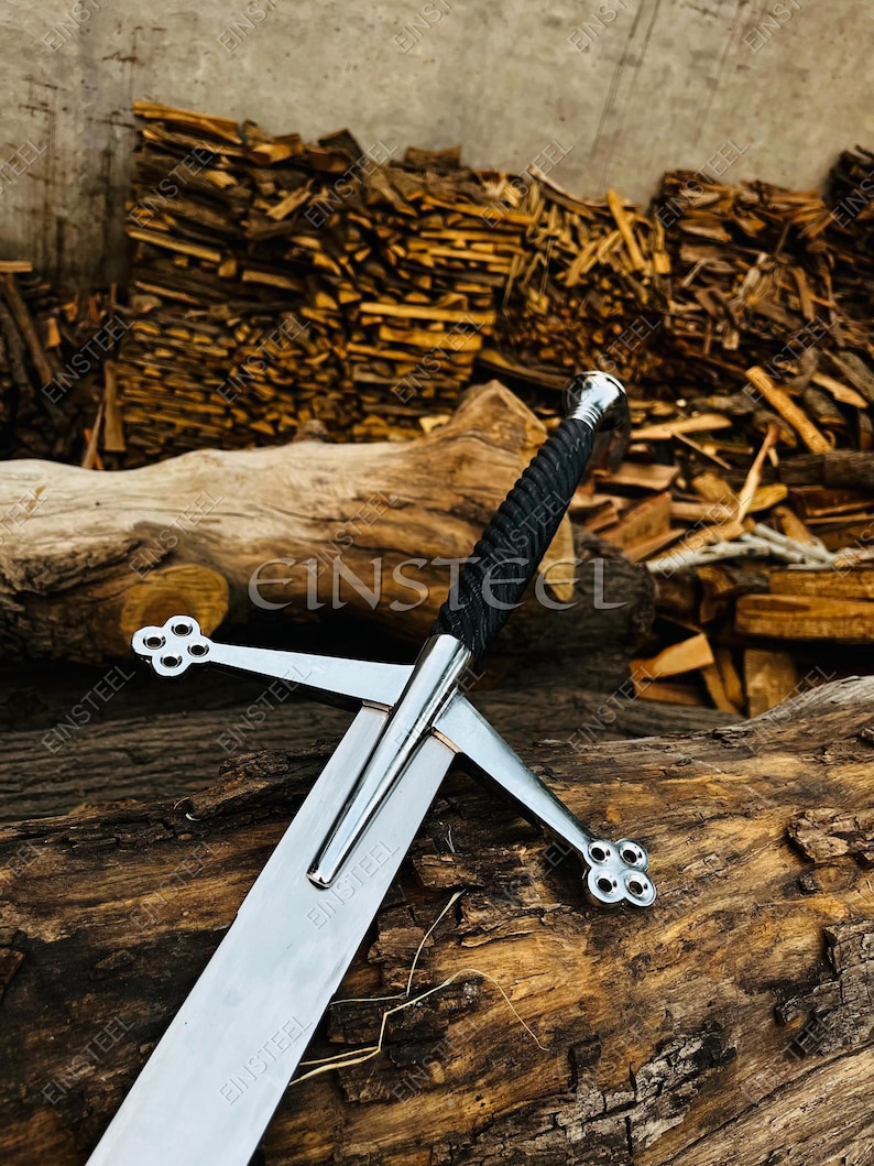 May include: A medieval-style sword with a polished steel blade and crossguard. The handle is wrapped in black material, and the pommel is silver. The sword rests on a weathered wooden surface, with a background of stacked firewood.