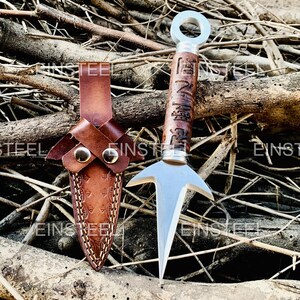 Op de afbeelding: Een zilveren metalen kunai-mes met een bruine houten handgreep en een leren schede. De handgreep heeft gegraveerde Japanse karakters. De schede is van bruin leer met decoratieve stiksels en twee messing studs. Het mes wordt getoond op een bed van twijgen.
