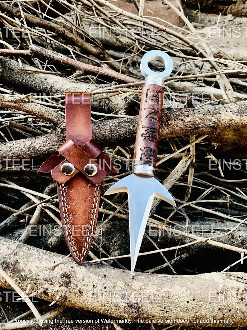 Op de afbeelding: Een zilveren metalen kunai-mes met een bruine houten handgreep en een leren schede. De handgreep heeft gegraveerde Japanse karakters. De schede is van bruin leer met decoratieve stiksels en twee messing studs. Het mes wordt getoond op een bed van twijgen.
