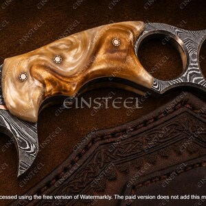 Pu&ograve; includere: Un coltello karambit con una lama in acciaio Damasco a motivi e un manico in resina lucida con vortici color marrone chiaro e crema. Il manico &egrave; fissato con tre rivetti metallici circolari e include un grande anello per il dito.