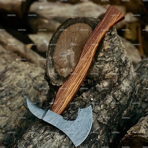 Könnte beinhalten: Eine handgefertigte Axt mit einem gebogenen Holzgriff und einer gemusterten Stahlklinge. Die Axt liegt auf einer verwitterten Holzoberfläche und zeigt ihre detaillierte Handwerkskunst. Die Klinge hat ein einzigartiges, strukturiertes Design.