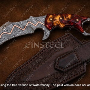 Pu&ograve; includere: Un coltello karambit curvo con lama in acciaio damasco caratterizzata da una linea a zigzag in rame intarsiato. L'impugnatura &egrave; in resina marmorizzata rossa e ambra con tre perni in ottone e un anello per il dito sul pomolo. Fodero incluso.
