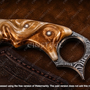 Pu&ograve; includere: Un coltello karambit con un manico dal motivo marmorizzato marrone e crema, fissato da due rivetti metallici circolari. La lama presenta un distinto motivo in acciaio damasco e include un prominente anello per le dita alla base.