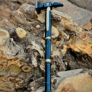 May include: A black and gold hammer with a decorative design. The hammerhead features a skull emblem. The handle is a dark blue color with gold accents. The hammer is displayed against a backdrop of wood.
