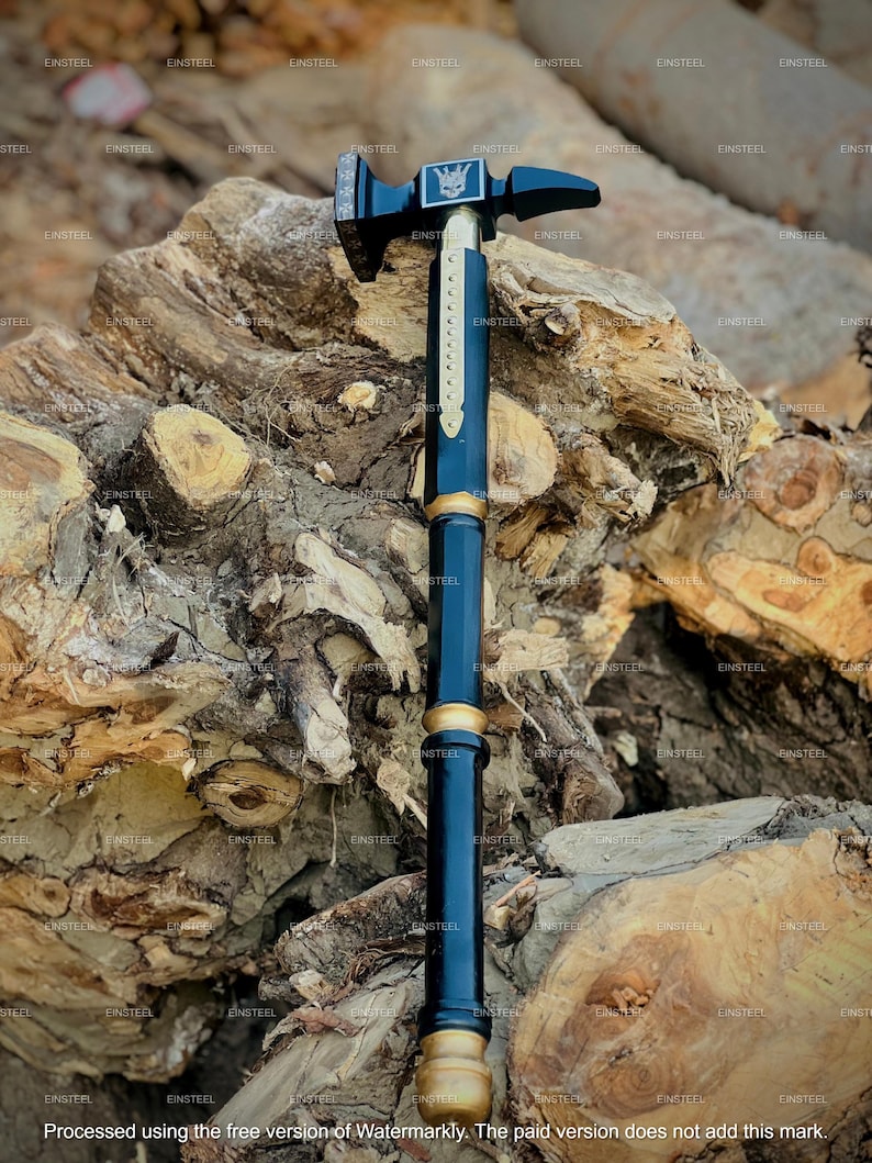 May include: A black and gold hammer with a decorative design. The hammerhead features a skull emblem. The handle is a dark blue color with gold accents. The hammer is displayed against a backdrop of wood.
