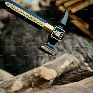 May include: A black and gold hammer with a decorative handle and head. The hammer features a gold emblem and is positioned over a background of wooden logs. The handle is adorned with gold accents and rivets.
