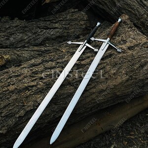 May include: Two long, silver swords with cross-shaped hilts and pointed blades rest on a dark wooden surface. One sword has a black handle, and the other has a brown, wrapped handle. The word "EINSTEEL" is visible on the blades.