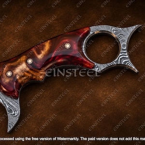 Pu&ograve; includere: Un coltello karambit caratterizzato da una lama in acciaio Damasco con un distinto motivo ondulato e un manico in resina lucida rossa e marrone. Il manico &egrave; fissato con tre rivetti in ottone e include un anello circolare per il dito sul pomolo.