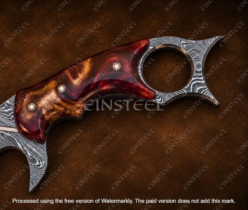 Pu&ograve; includere: Un coltello karambit caratterizzato da una lama in acciaio Damasco con un distinto motivo ondulato e un manico in resina lucida rossa e marrone. Il manico &egrave; fissato con tre rivetti in ottone e include un anello circolare per il dito sul pomolo.