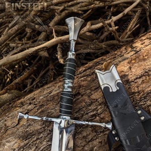 Puede incluir: Una espada de metal plateado con empuñadura envuelta en cuero negro y una vaina de cuero negro a juego. La espada presenta una hoja de acero pulido, una guarda con diseño retorcido y un pomo facetado. La vaina tiene detalles en metal plateado.