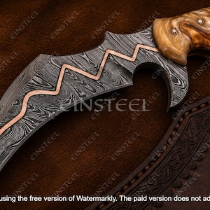 Pu&ograve; includere: Un coltello a lama curva con un motivo in acciaio Damasco scuro e un intarsio in rame a zigzag lungo il dorso. Il manico &egrave; realizzato in materiale marmorizzato marrone chiaro con piccoli rivetti metallici. &Egrave; visibile un fodero in pelle.